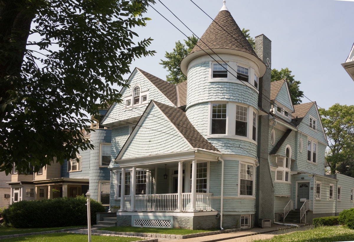 The exterior of a historic blue Victorian home is presented, showcasing intricate shingle patterns and a prominent turret. A wraparound porch with white railings invites relaxation, while well-maintained landscaping and trees provide a shaded, serene setting.
