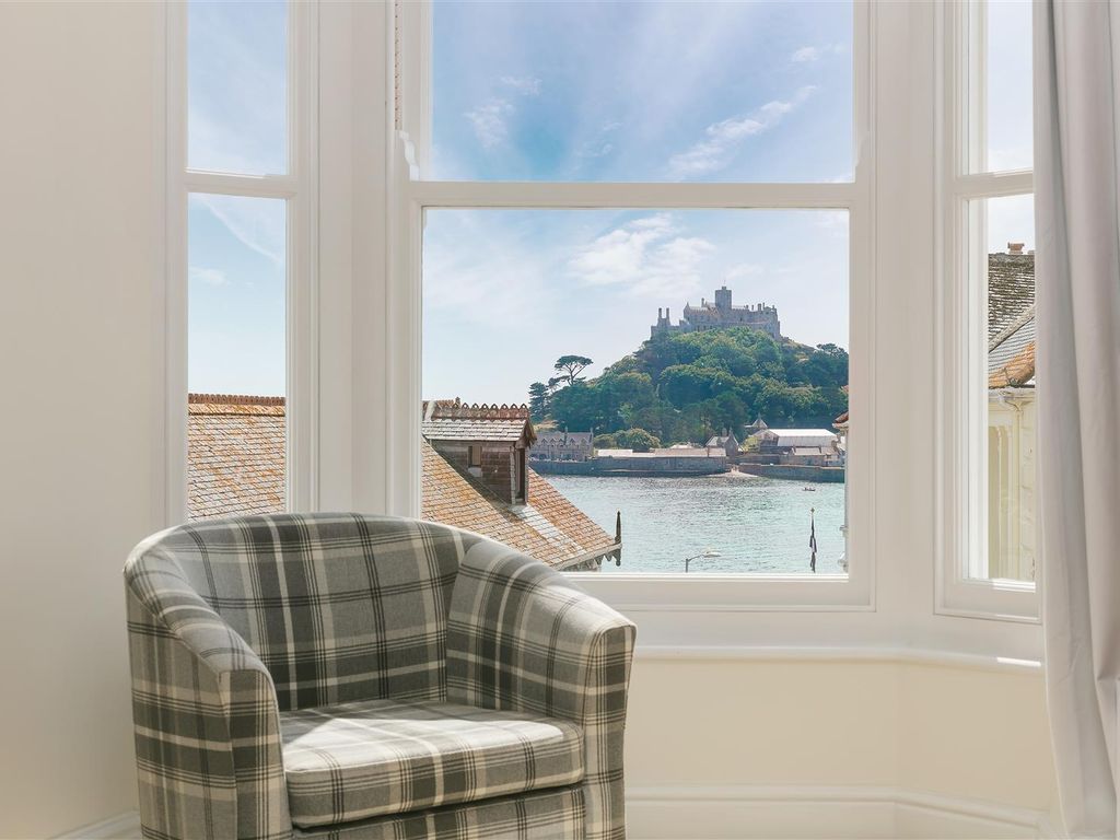 A comfortable armchair with a checkered pattern is positioned near a window, offering a clear view of St Michael's Mount and the water below. Natural light fills the space, highlighting the scenic backdrop of the castle atop the island.