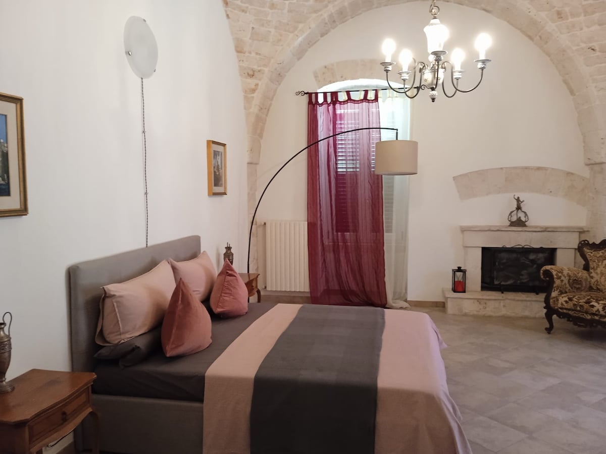 A spacious bedroom features a comfortable bed with a soft cover and decorative pillows. An elegant chandelier hangs from a curved ceiling. Soft drapes frame a window, and a vintage armchair adds character to the room. A fireplace suggests warmth and charm.