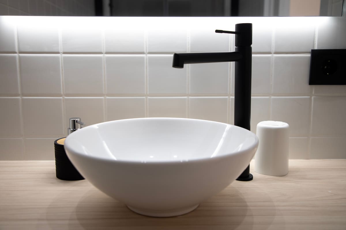 A modern bathroom sink is positioned on a light wooden countertop. A round white bowl sink is paired with a sleek black faucet. Nearby, a soap dispenser and a cylindrical container are placed, providing essential amenities. Soft lighting illuminates the white tiled backsplash.