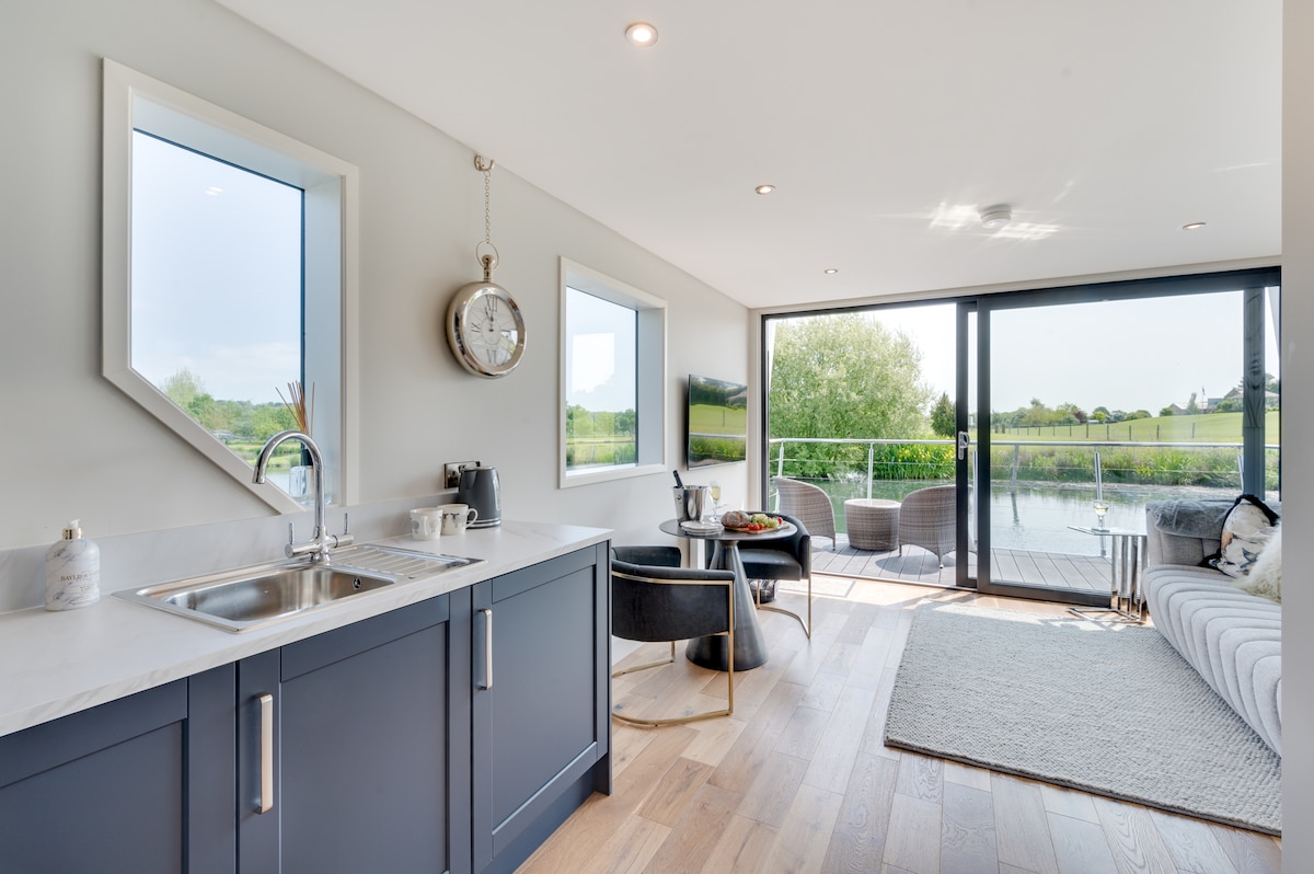 An open-plan living area features large windows that fill the space with natural light. A modern kitchen with sleek cabinetry is complemented by stylish seating options, including a comfortable sofa and armchairs. A sliding door leads to an outdoor deck with views of the surrounding landscape.
