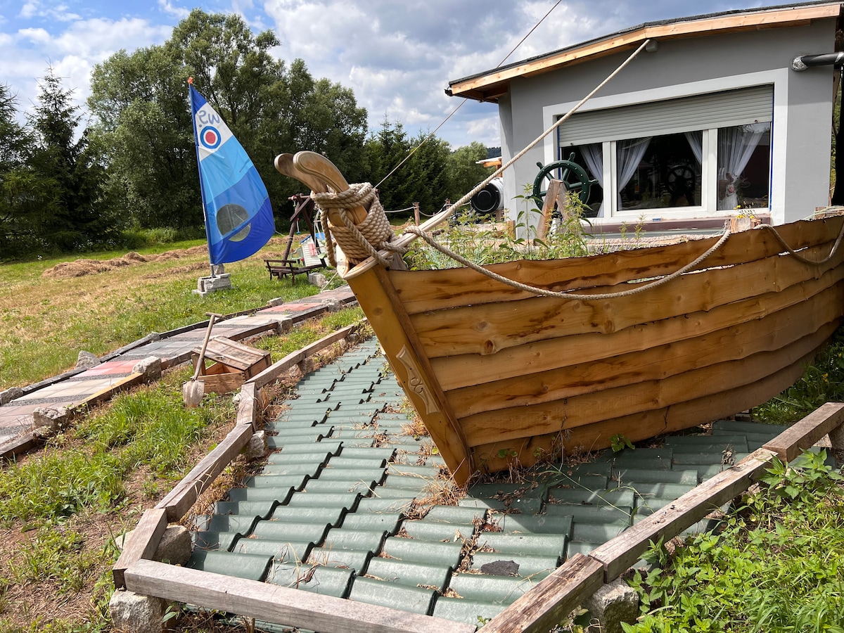 A wooden boat is placed on a grassy area, with a sloped green roof visible underneath. A small blue sail is seen in the background, adding a touch of color to the natural setting. Trees are present in the distance, enhancing the landscape.