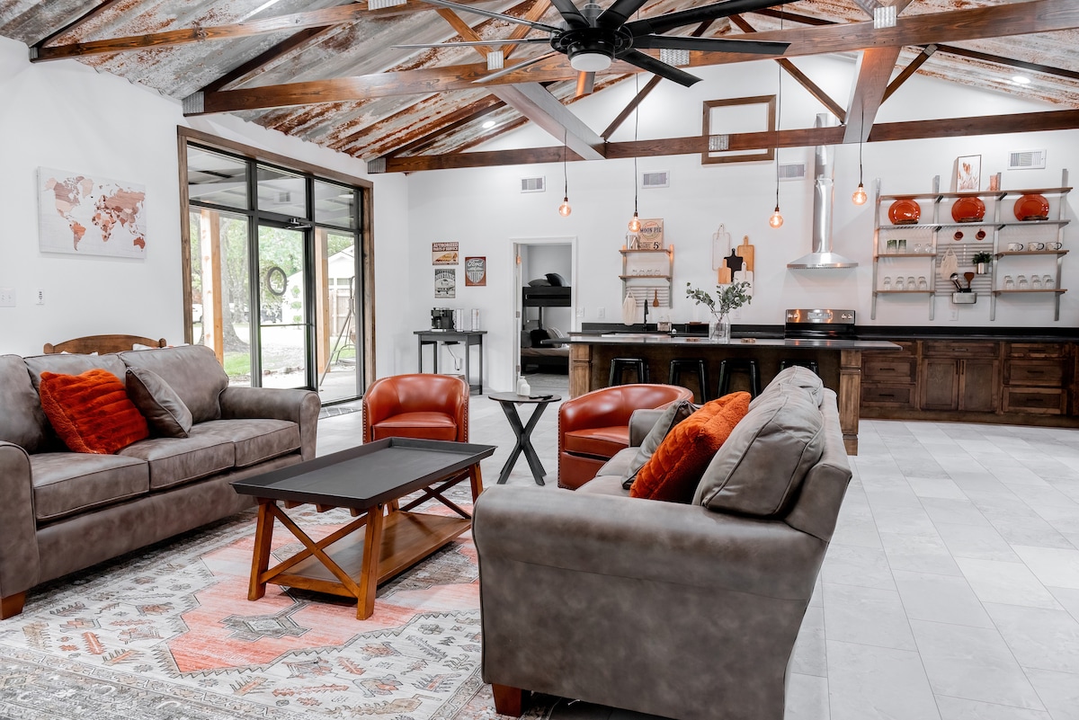 A spacious living area features a rustic ceiling with exposed beams. Comfortable sofas with orange accents are arranged around a dark coffee table, complemented by armchairs. The open concept leads to a kitchen area with wooden cabinetry and decor, illuminated by pendant lights.