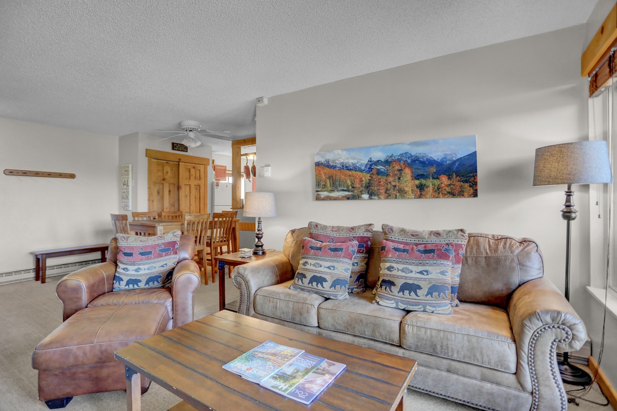 The living room features a comfortable leather sofa accented by colorful throw pillows. A wooden coffee table sits in front, displaying brochures. Natural light filters in through large windows, highlighting a scenic mural of mountains and trees in autumn colors.