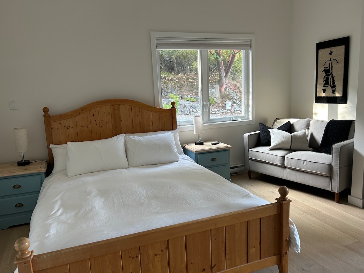 A spacious bedroom features a wooden bed with white bedding and a cozy sitting area with a gray couch. Two bedside tables with lamps complement the room. A large window allows natural light and offers a view of the surrounding greenery.