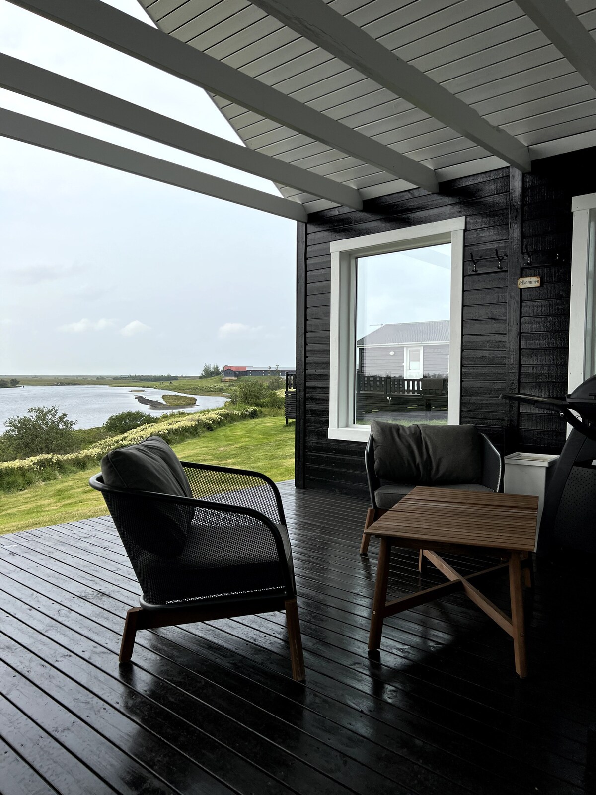 A covered outdoor seating area is visible, featuring two comfortable chairs and a small wooden table. Large windows frame views of the grassy landscape and the winding Rangá River, enhancing the connection to nature.