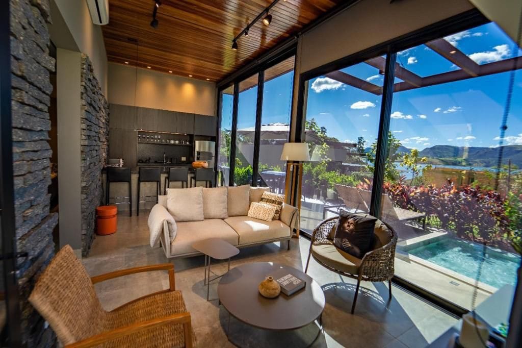 A modern living area features large glass doors offering views of the outdoor space. Comfortable seating includes a light-colored sofa, a round table, and woven chairs. A sleek kitchenette is visible, surrounded by a blend of natural stone and wood elements, enhancing the overall contemporary design.