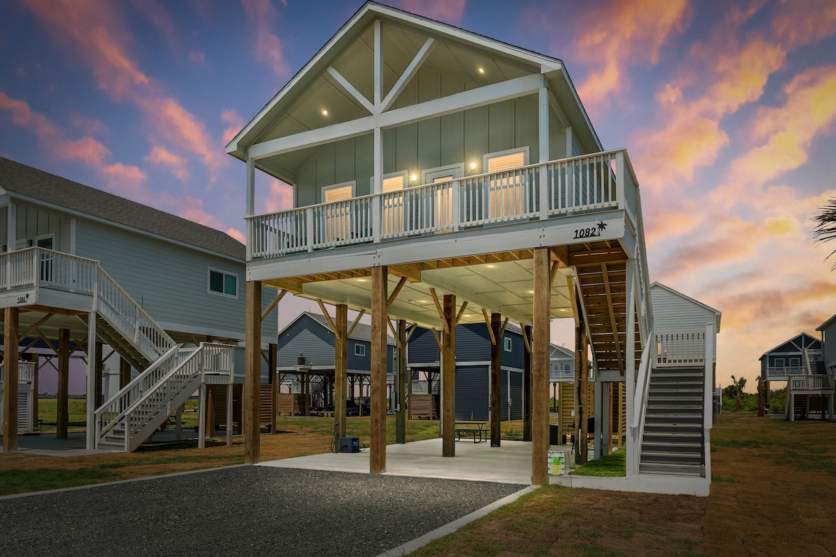 A modern beach house on stilts is showcased against a colorful sunset. The elevated structure features a spacious deck with multiple glass doors. Ample parking space is visible below, surrounded by grassy areas. Nearby homes can be seen in the background, adding to the coastal atmosphere.