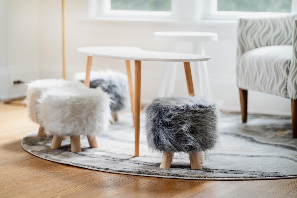 A stylish seating area features a low white table complemented by two cozy ottomans covered in soft, textured fur. Natural wood legs support the ottomans, providing a touch of warmth to the inviting space. A patterned area rug enhances the overall aesthetic.