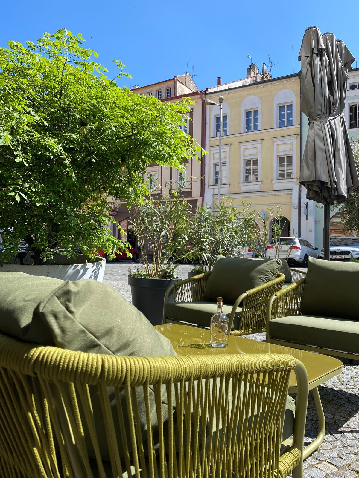 A cozy outdoor seating area features a green wicker sofa with plush cushions, surrounded by potted plants. The backdrop includes colorful buildings under a clear blue sky, creating a welcoming space for relaxation.