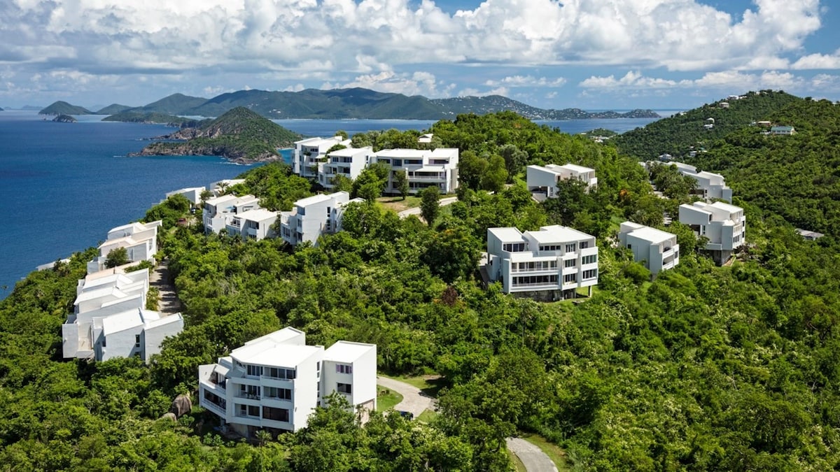 Aerial view of a hillside community featuring modern white buildings surrounded by lush green vegetation and overlooking the ocean. The landscape showcases the combination of nature and contemporary architecture, with distant hills and cloudy skies adding depth to the scene.
