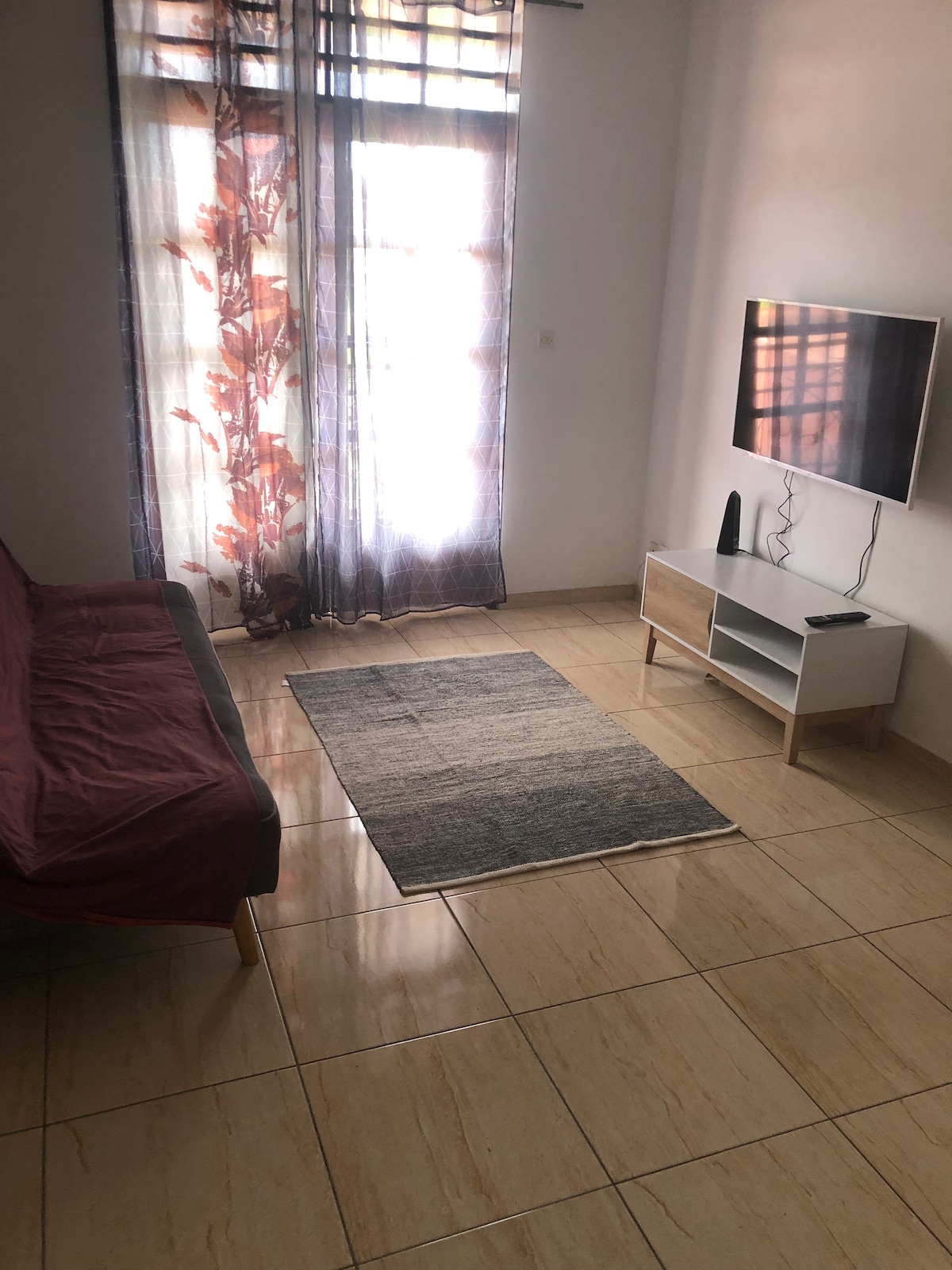 A bright living area is shown with a large window covered by flowing curtains with floral patterns. A dark sofa is positioned against the wall, complemented by a simple rug on a shiny tiled floor. A television is mounted on a white unit nearby.