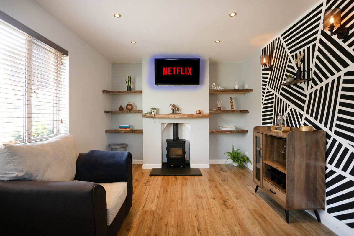 A modern living area features a comfortable sofa with a dark blue armrest. Wall shelves are adorned with decorative items, and a smart TV is mounted above a fireplace. A light wooden floor complements the geometric-patterned accent wall.