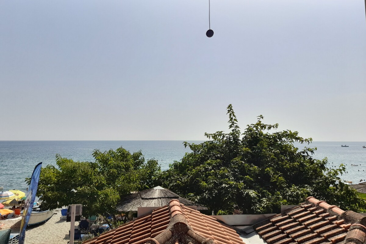 A serene view of the ocean is presented, framed by lush green trees and terracotta rooftops. The calm sea stretches toward the horizon under a clear sky, inviting a sense of tranquility. Subtle details of beach setups can be glimpsed in the distance.
