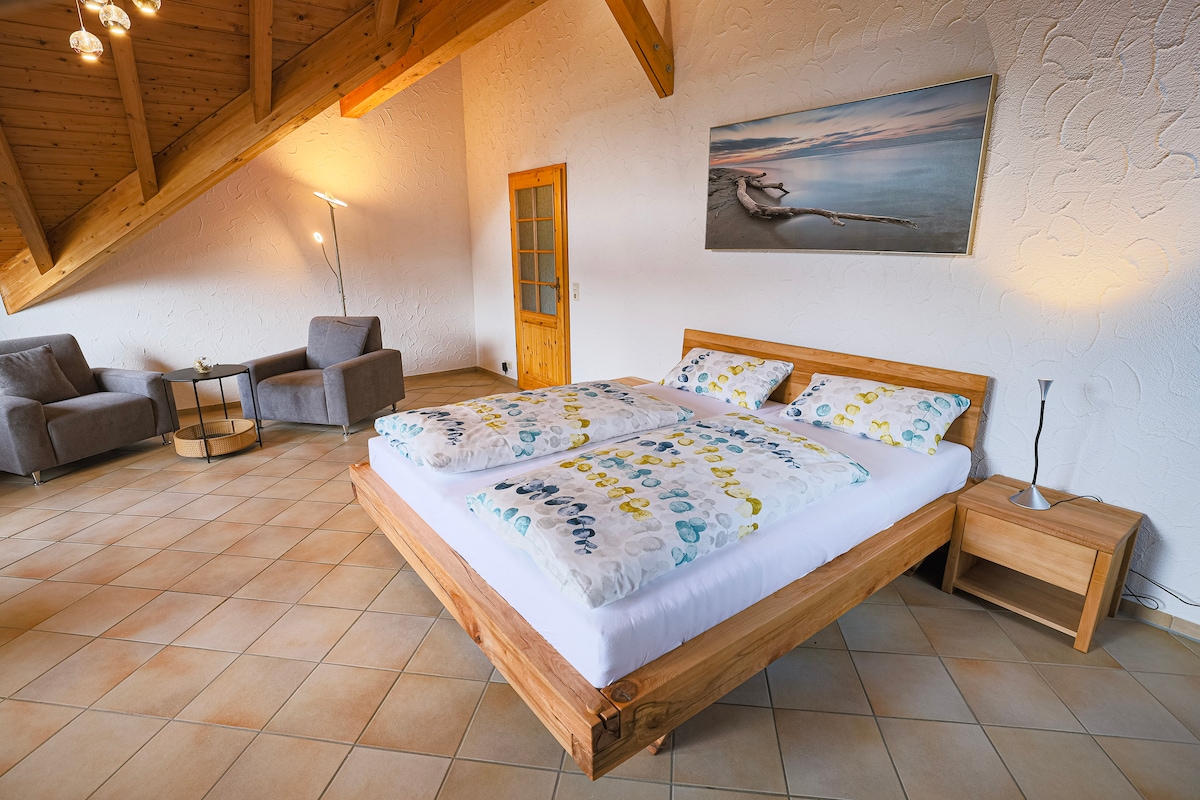 A spacious sleeping area features a wooden bed with colorful patterned linens. Soft seating options are positioned nearby, along with a decorative table. Natural light fills the room, highlighting the textured wall and wooden beams of the ceiling.