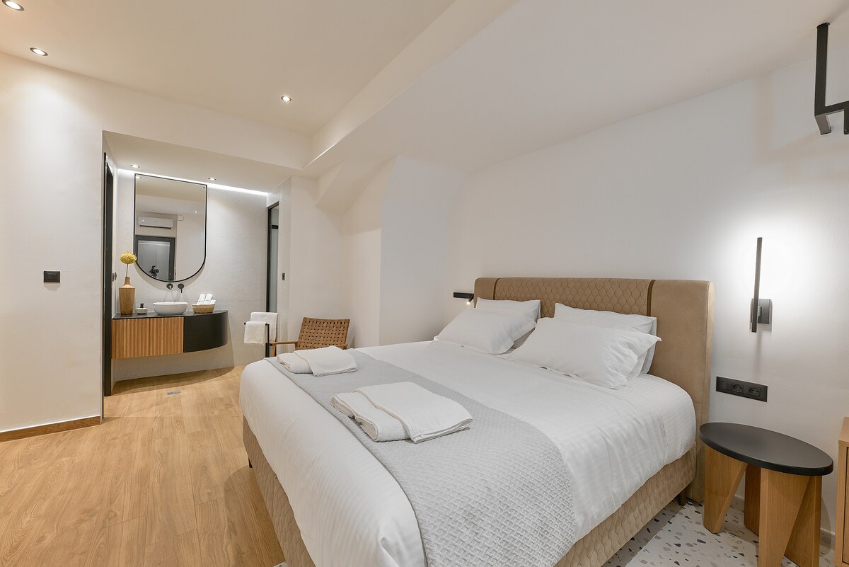 A spacious bedroom features a comfortable king bed dressed with quality linens. Natural light enters through a nearby window, illuminating the simple, modern decor. An en suite bathroom is visible in the background, equipped with contemporary fixtures and finishes.