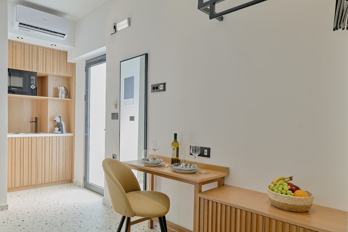 A clean and modern dining area is presented, featuring a light-colored wooden table set for two. A wall-mounted mirror and a basket of fruits are visible, alongside kitchen appliances in the background, enhancing the apartment's functionality and inviting ambiance.
