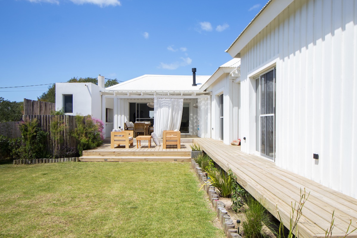 A spacious outdoor area features a wooden deck with seating arrangements and a dining table. The white exterior of the house complements the lush green lawn, while a path lined with plants leads toward the entrance. Sunlight illuminates the scene under a clear blue sky.
