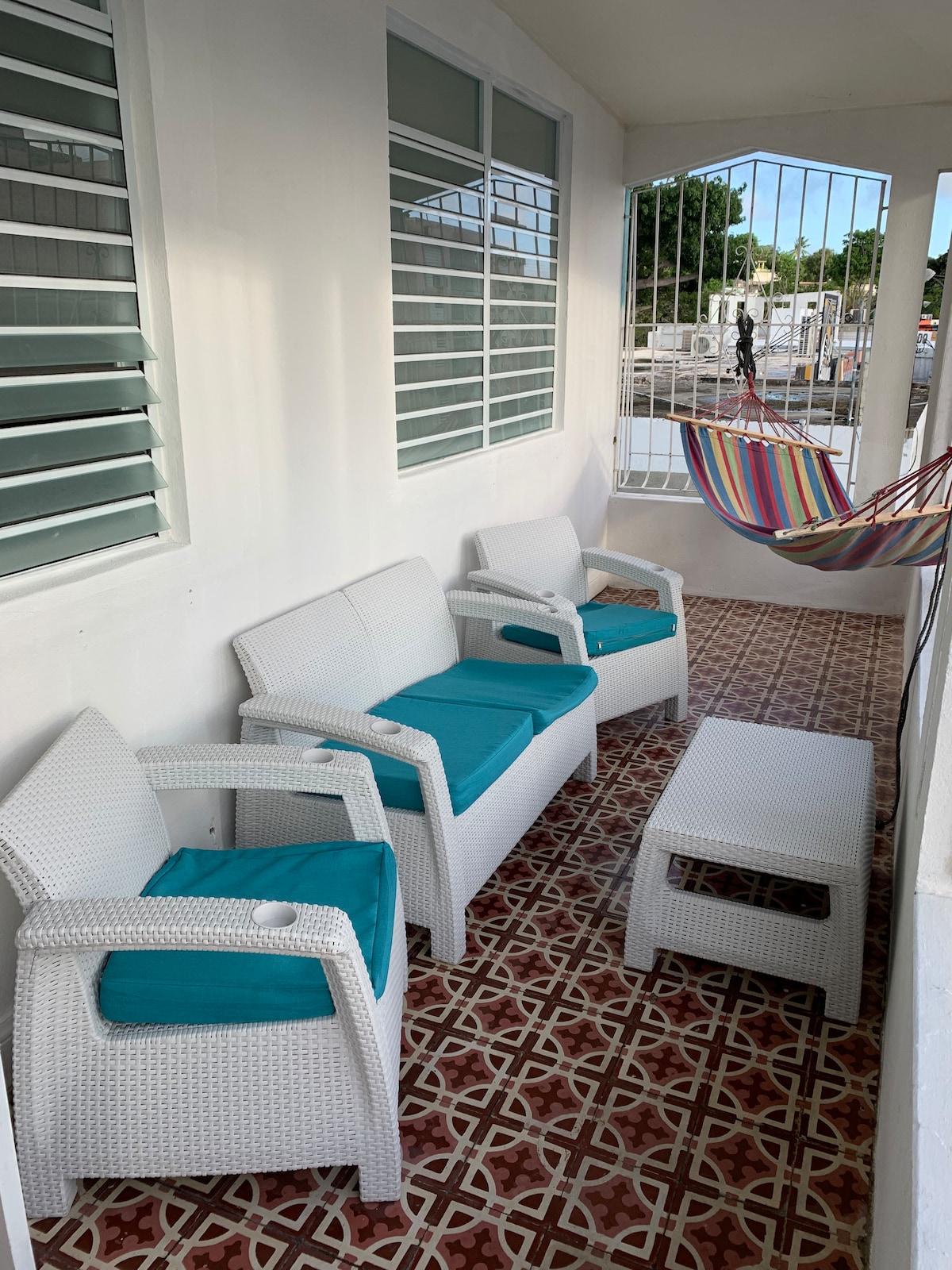 A spacious balcony is furnished with four white wicker chairs featuring turquoise cushions and a rectangular coffee table. A vibrant, multicolored hammock is visible, suspended nearby. The patterned floor tiles add a decorative touch, while the surrounding railings allow for fresh air and light.