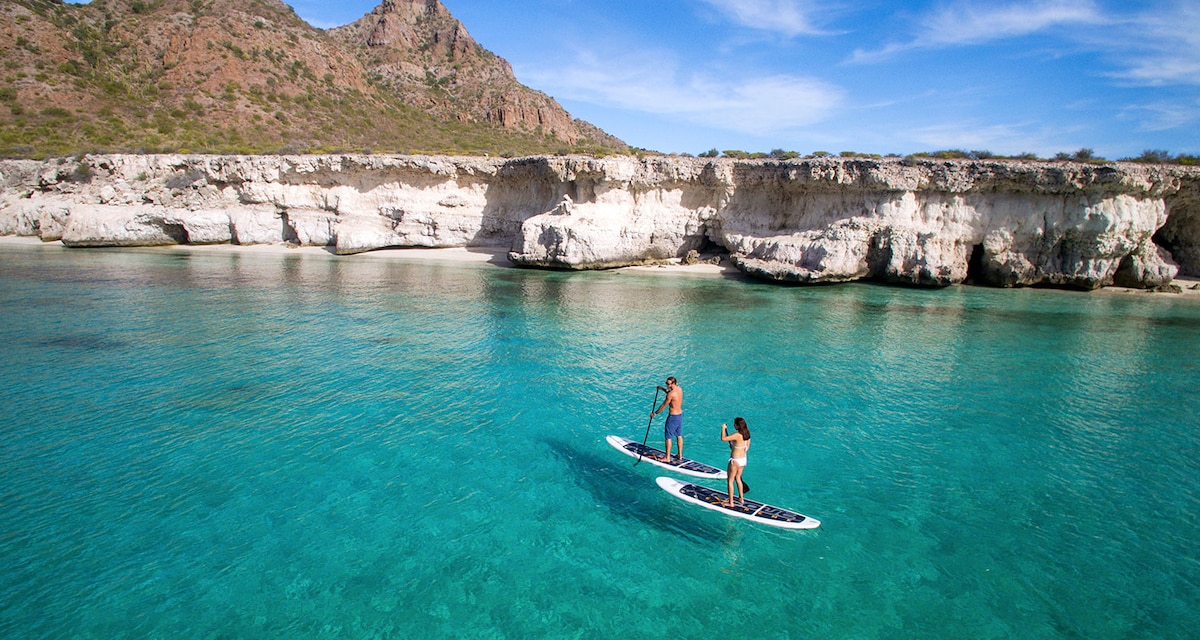 Two individuals are seen paddleboarding on clear turquoise waters, surrounded by rocky cliffs and a serene coastline. The sun illuminates the scene, capturing the peaceful atmosphere of the area with gentle waves lapping against the boards.
