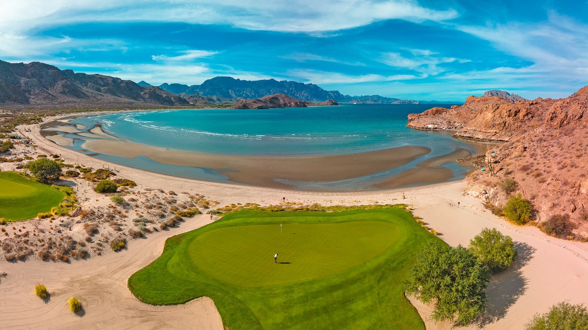 A panoramic view of a golf course foregrounded by a vibrant green fairway, leading to a serene beachside. The calm waters of the bay reflect the distant mountains, while a clear blue sky frames the scene, highlighting the natural beauty of the untouched landscape.