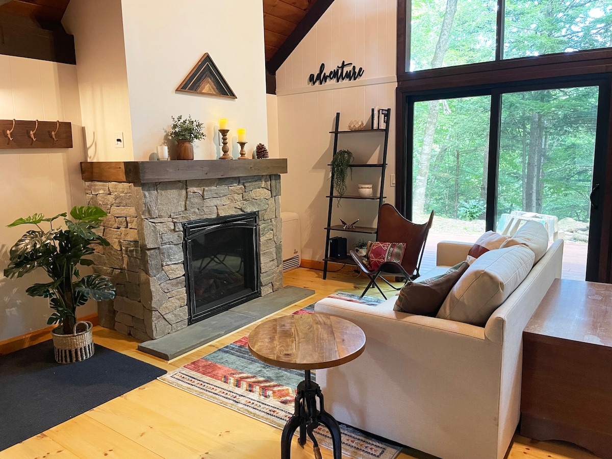 A cozy living area features a natural stone fireplace framed by a wooden mantle. A light-colored sofa, complemented by a patterned area rug, faces a large glass door that opens to the outdoor space. A small side table and a plant add to the inviting ambiance.