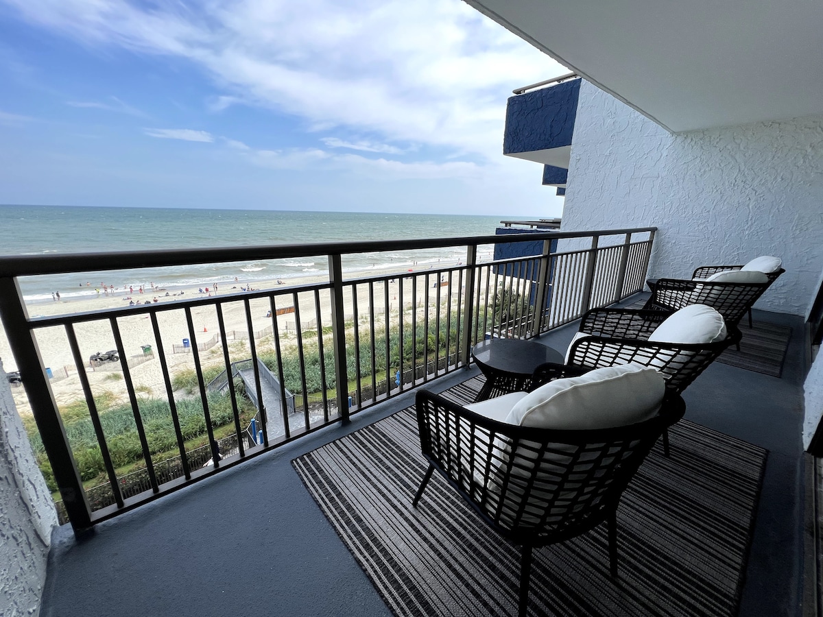 A private oceanfront balcony features comfortable seating for four, overlooking the sandy beach and ocean waves. The flooring is finished with a stylish rug, and the railing provides an unobstructed view of the shoreline and horizon.