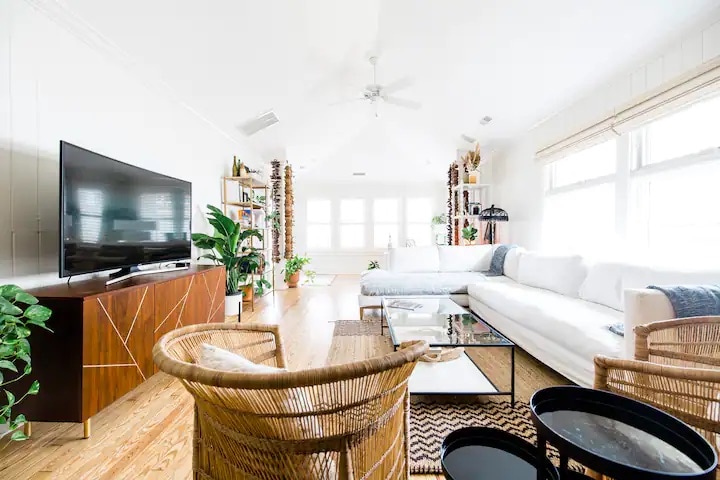 A bright living space features large windows allowing natural light to illuminate the room. A spacious white sectional sofa is positioned near a glass coffee table, surrounded by lush green plants. A flat-screen television is mounted on a wooden console, adding a modern touch.