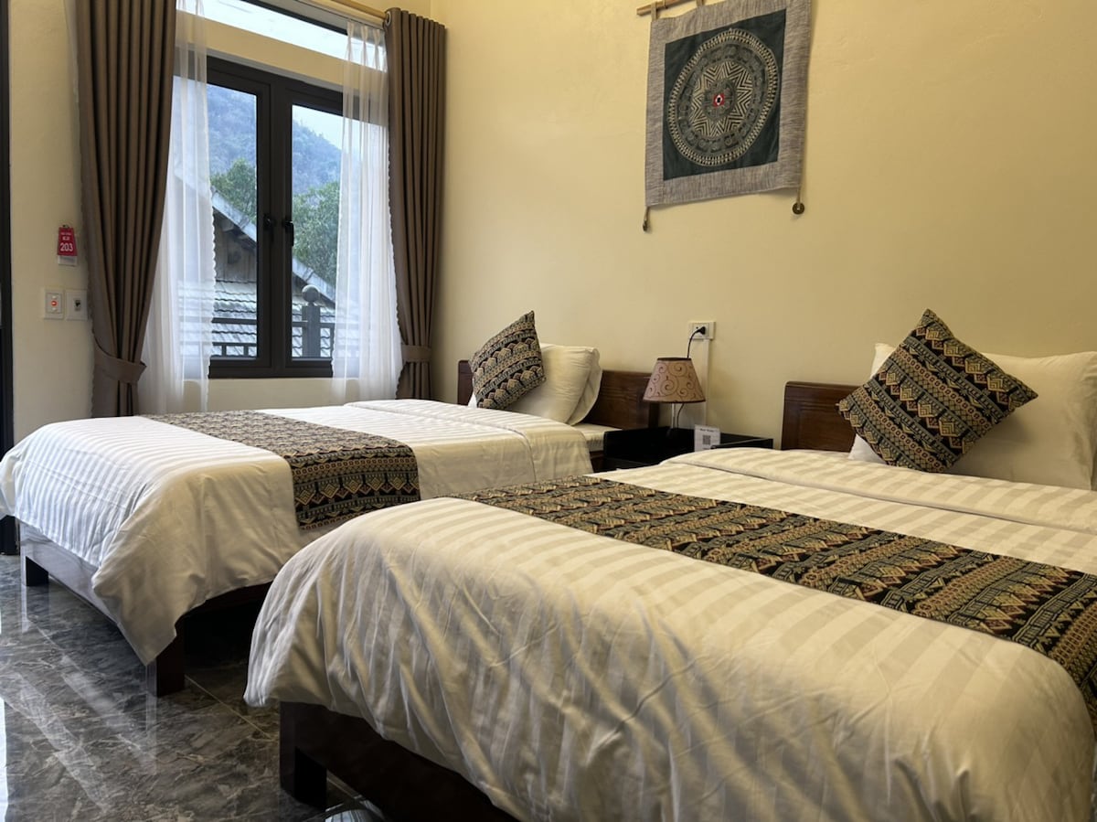A well-lit room features two twin beds, each dressed in crisp white linens with patterned throws. The room includes decorative pillows and a bedside lamp on a dark wood nightstand. Large windows allow natural light to fill the space, complemented by soft curtains.