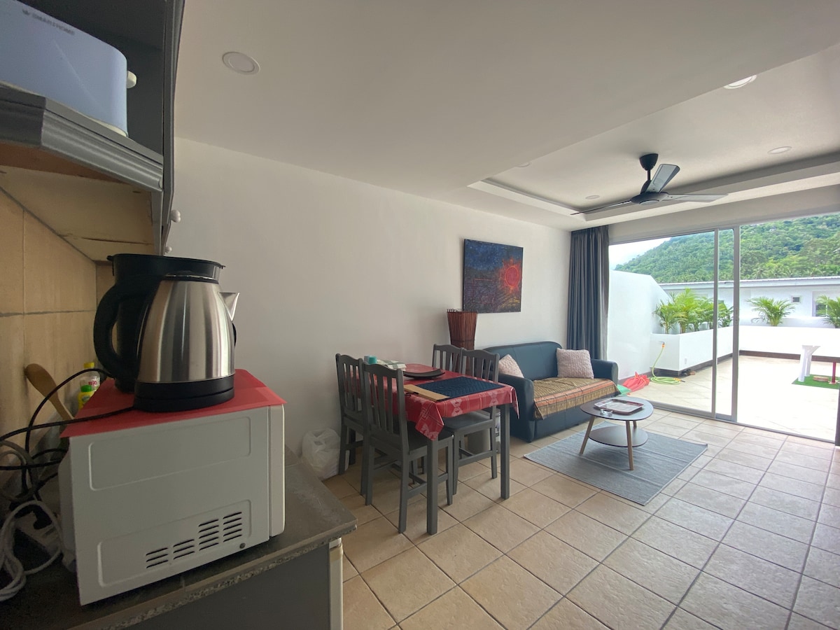 The living area features a cozy couch facing a large glass door that opens to a balcony. A dining table with four chairs is set nearby, and the kitchen area is equipped with a microwave and electric kettle, complemented by tiled flooring and soft natural light.