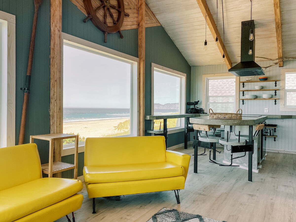 A bright living space features two yellow sofas and a dining area with a large table. Expansive windows provide views of the beach and ocean. A modern kitchen area is equipped with a central island, dark overhead lighting, and open shelves.