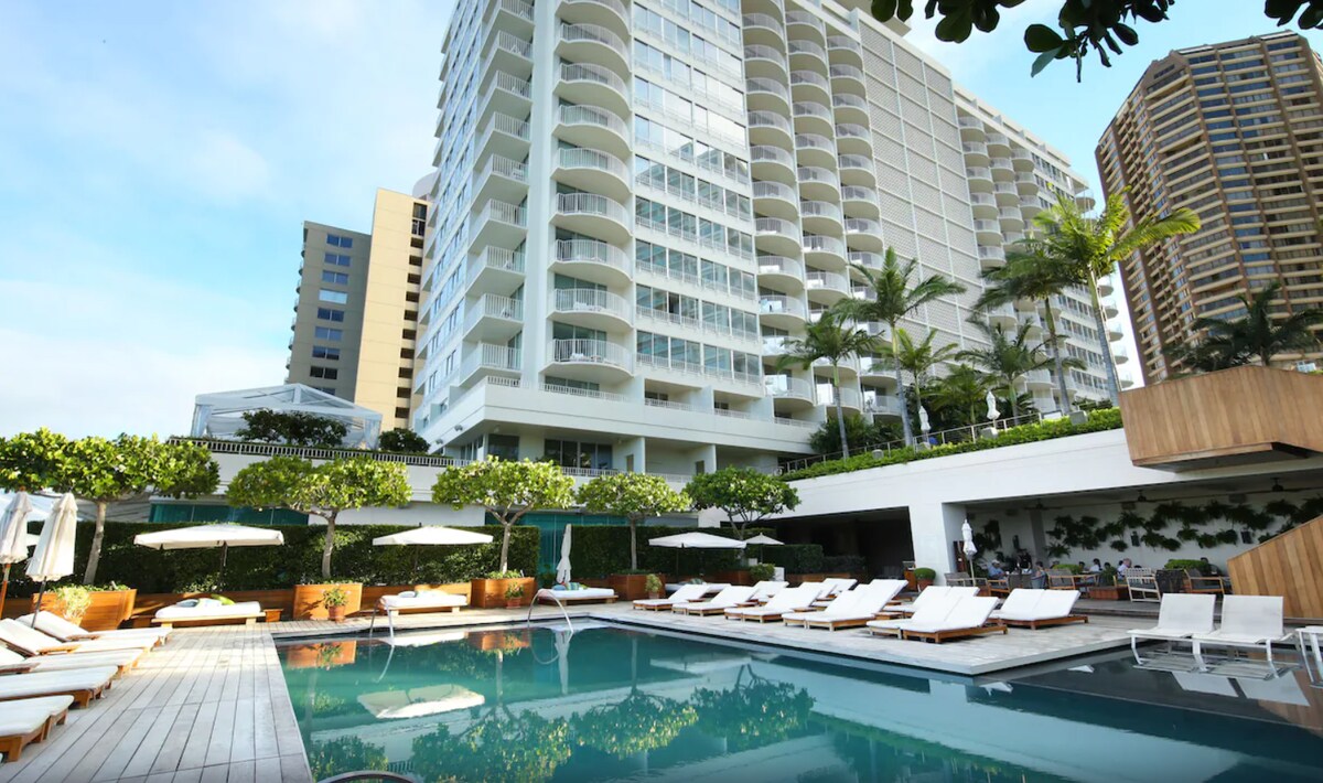 A modern resort pool area features a spacious swimming pool surrounded by lounge chairs. The building's sleek facade is complemented by lush greenery and palm trees, creating a refreshing atmosphere. Sun umbrellas are positioned along the deck for shade.
