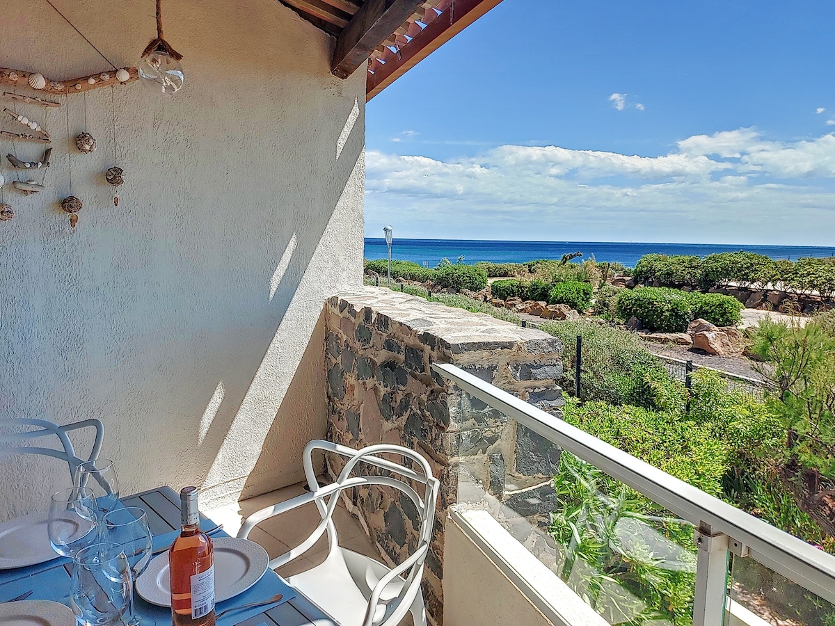 A terrace is showcased with a view of the sea, surrounded by greenery. A dining table set for four with plates, glasses, and a bottle of wine is visible. Natural light illuminates the space, complemented by a stone wall feature.
