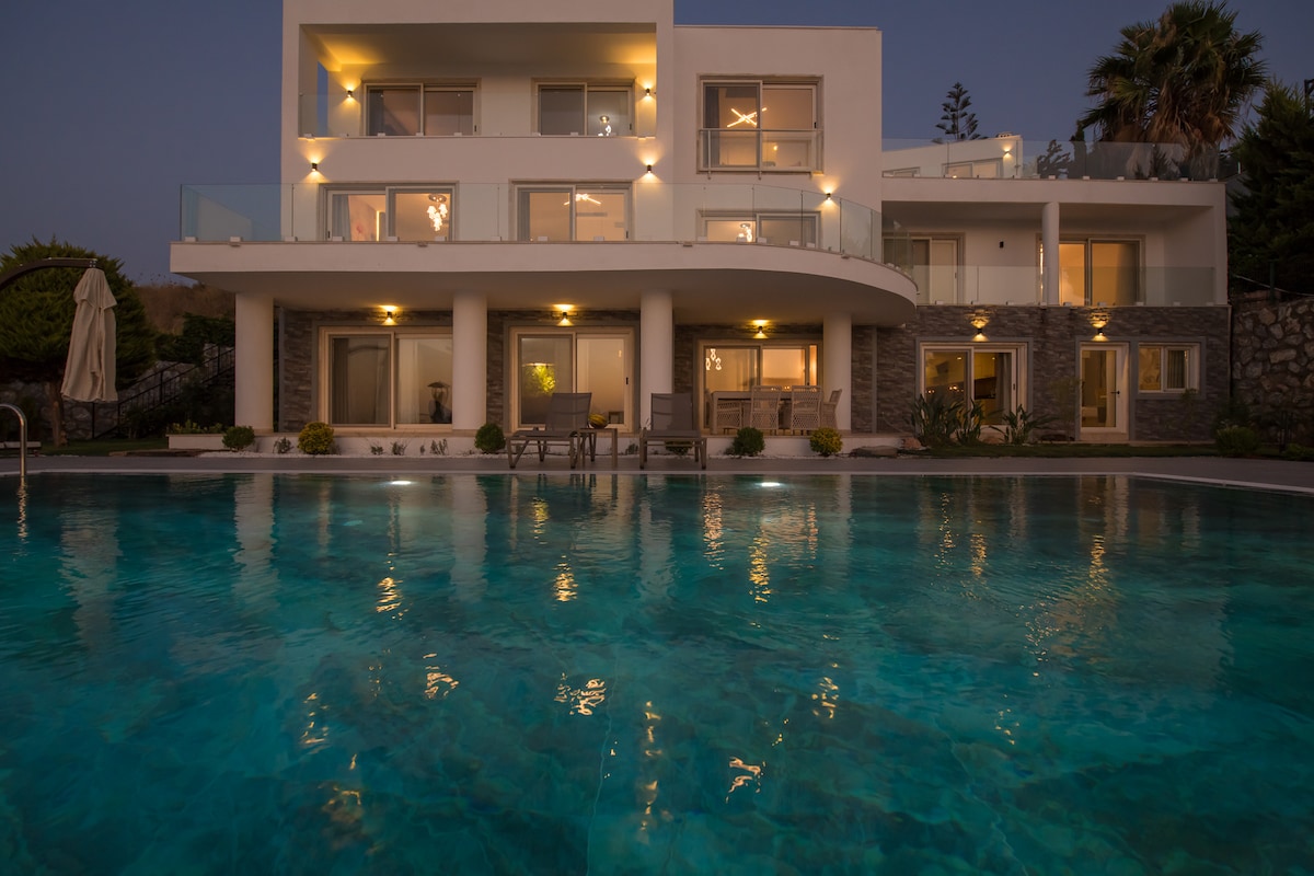 The villa is illuminated at dusk, reflecting off the tranquil swimming pool. Large glass doors lead to outdoor seating areas surrounded by well-maintained gardens. The multi-level structure is accented by modern architectural features and large terraces overlooking the pool.
