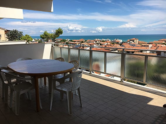 A spacious covered terrace features a round table surrounded by several white chairs. The railing offers a panoramic view of the sea and the surrounding area, with rooftops visible in the distance. Sunlight casts a soft shadow across the tiled floor.