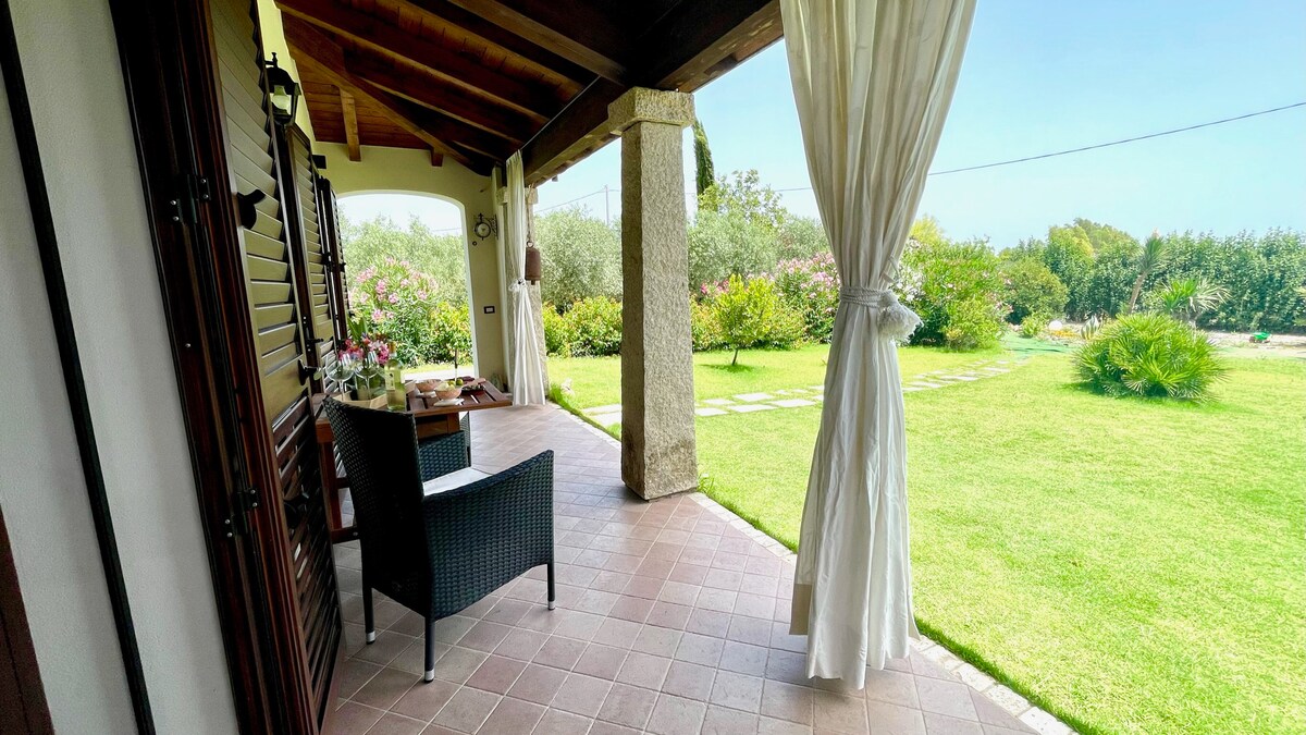A covered porch features a dark wicker chair beside a small table, ideal for outdoor relaxation. The tiled floor extends towards a lush garden, showcasing manicured grass and various plants in the background, under a clear blue sky.