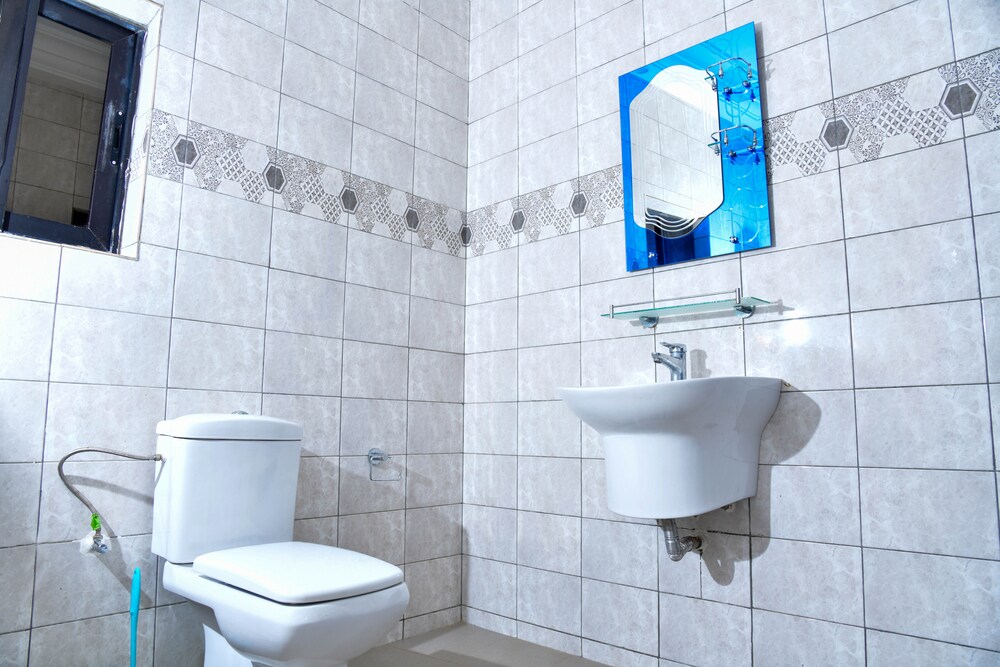 A clean and modern bathroom is presented, featuring light-colored tiled walls. A compact toilet is positioned next to a wall-mounted sink, which includes a reflective mirror above it. Natural light is filtered through a small window, enhancing the overall brightness of the space.