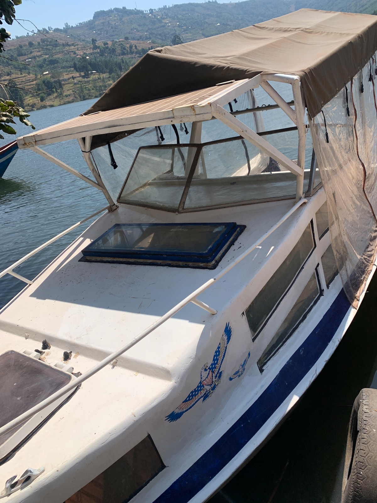 A white boat with a blue stripe is docked beside calm water. The boat features a covered seating area with transparent windows, allowing for visibility while providing shade. The serene landscape of green hills and trees can be seen in the background.