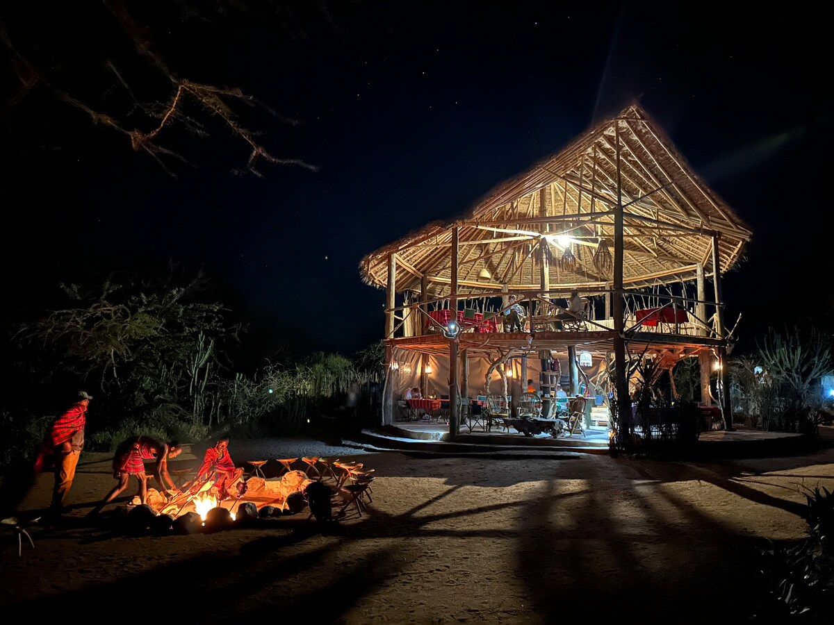 A traditional structure made of natural materials is featured against a night sky. The building is illuminated, revealing a spacious outdoor seating area. A campfire is positioned in the foreground, surrounded by individuals engaged in evening activities. Shadows cast by trees add depth to the scene.