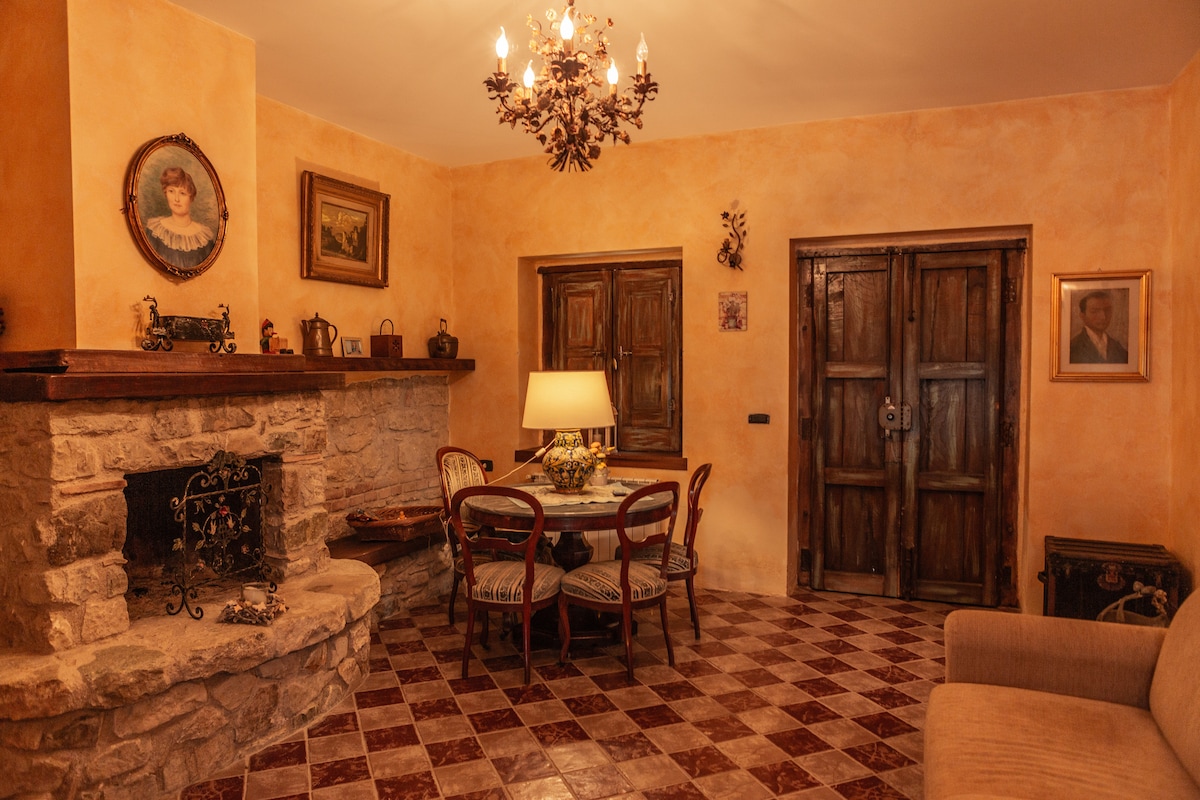 The cozy living area features a stone fireplace and a wooden dining table surrounded by four chairs. Natural light is provided by a lamp and a window, while warm, earthy tones adorn the walls. A soft sofa adds to the inviting atmosphere.