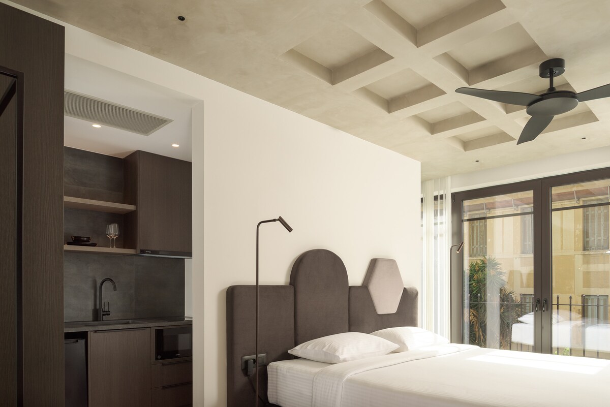 A contemporary bedroom features a spacious bed with a unique headboard design. A sleek kitchenette is visible in the background, complemented by modern cabinetry. Natural light fills the room through large windows with minimalistic treatments, enhancing the clean lines of the ceiling.