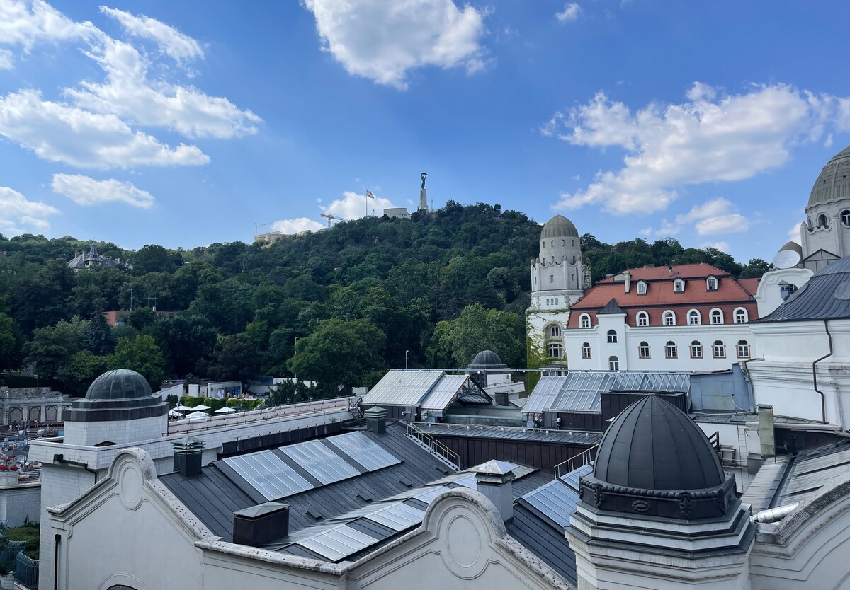 A panoramic view showcases rooftops of buildings with solar panels, framed by greenery and hills. Gellert Hill rises in the background, featuring a statue atop, while a partly cloudy blue sky adds depth to the scene.