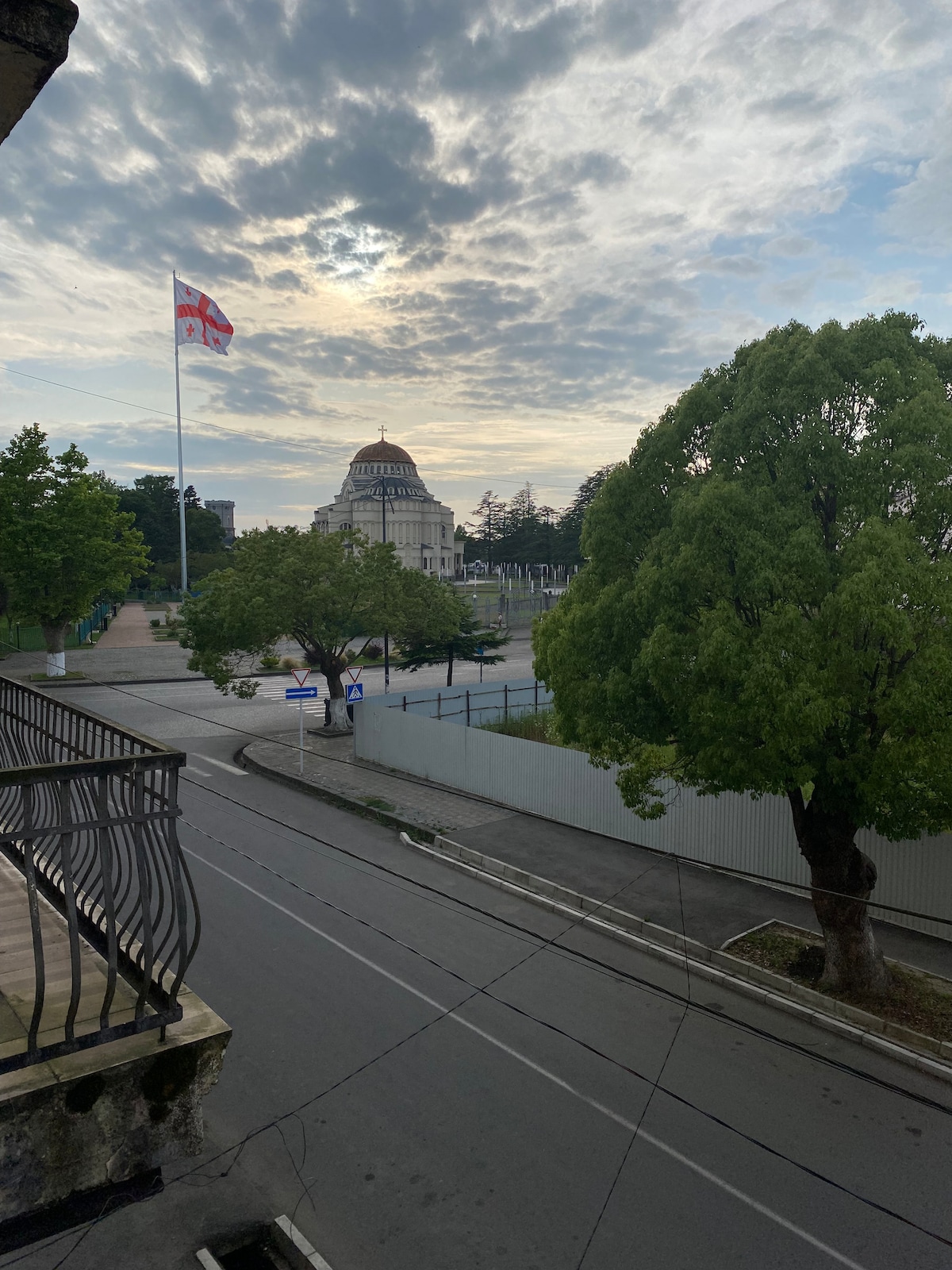 A view from a balcony captures a cityscape featuring a prominent building with a large dome. Lush green trees line the street, while a national flag flutters gently in the breeze against a cloudy sky.