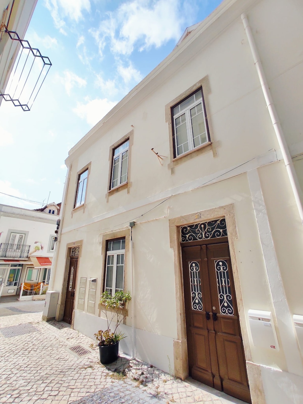 The exterior of a charming building is featured, showcasing traditional architectural details, including wooden doors and window panes. Bright sunlight illuminates the white facade, set on a cobblestone street lined with small shops, adding a touch of local character.
