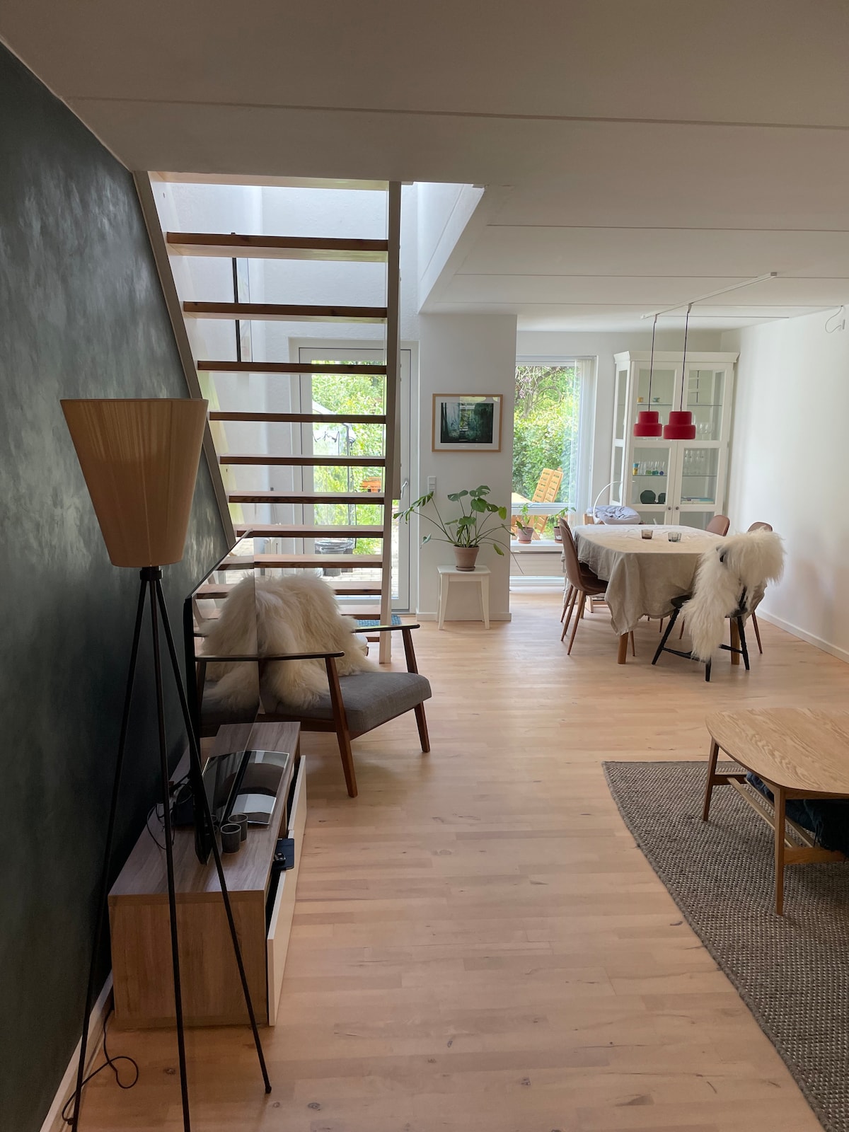 An open living space features a wooden staircase leading to the upper level. A comfortable armchair sits beside a low table, and a dining area is visible, adorned with a plant and cheerful light fixtures. Natural light streams in through large windows, illuminating the wooden flooring.