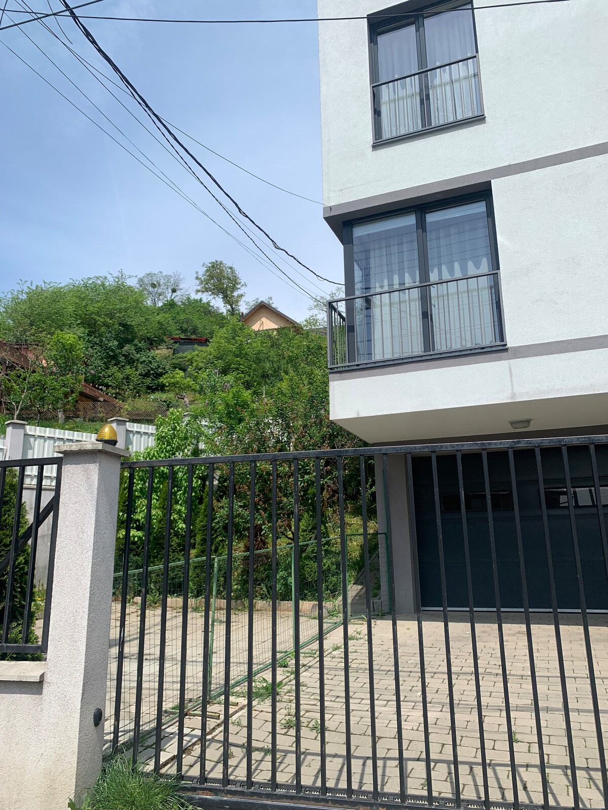 The image features a modern three-story building situated behind a black metal gate. A paved path leads to the entrance, with greenery visible in the surrounding area. The upper floor has large windows with sleek frames, allowing natural light to enter.