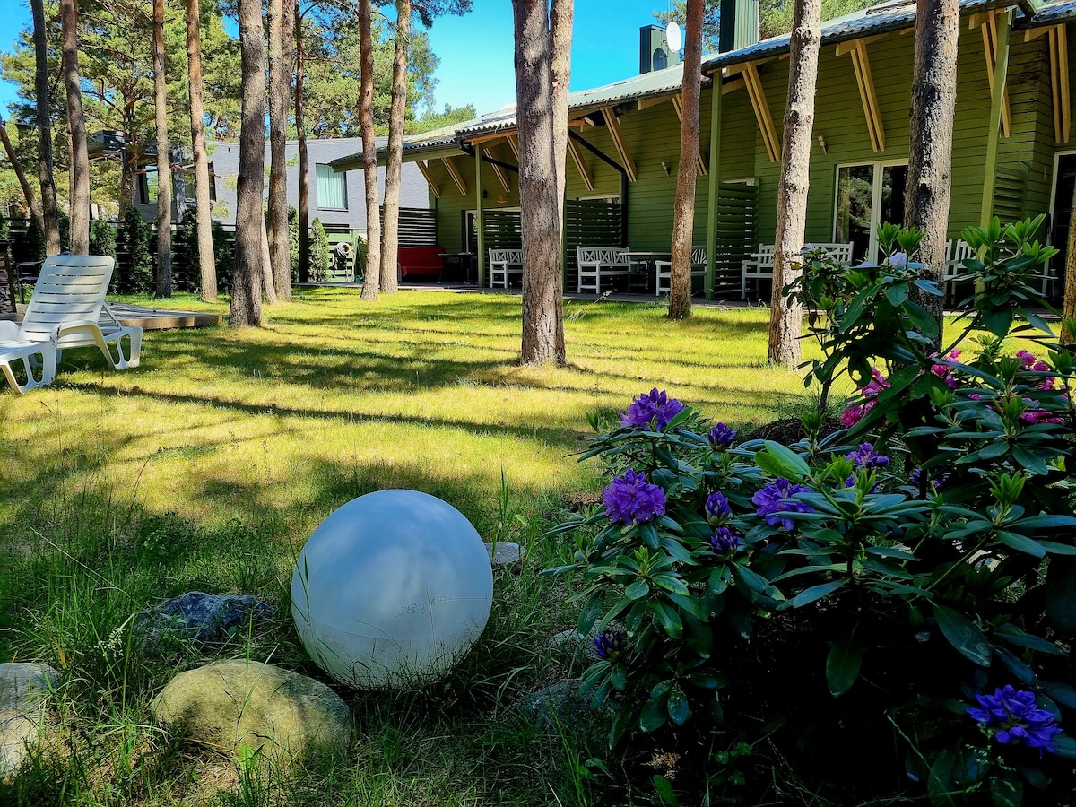 A serene outdoor space is presented with well-maintained grass and a variety of trees casting gentle shadows. Colorful flowers are featured in the foreground, along with a smooth white decorative stone. The spacious cottage with a green exterior is visible in the background.