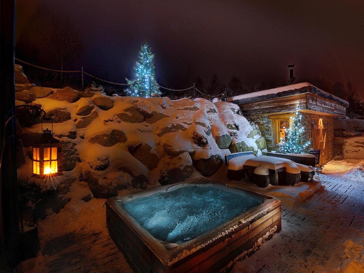 A hot tub sits invitingly on a wooden deck, surrounded by snow-covered rocks and illuminated by soft, glowing lights from nearby trees. A cozy cabin is positioned beside the hot tub, with warm light emanating from its windows against the wintry backdrop.
