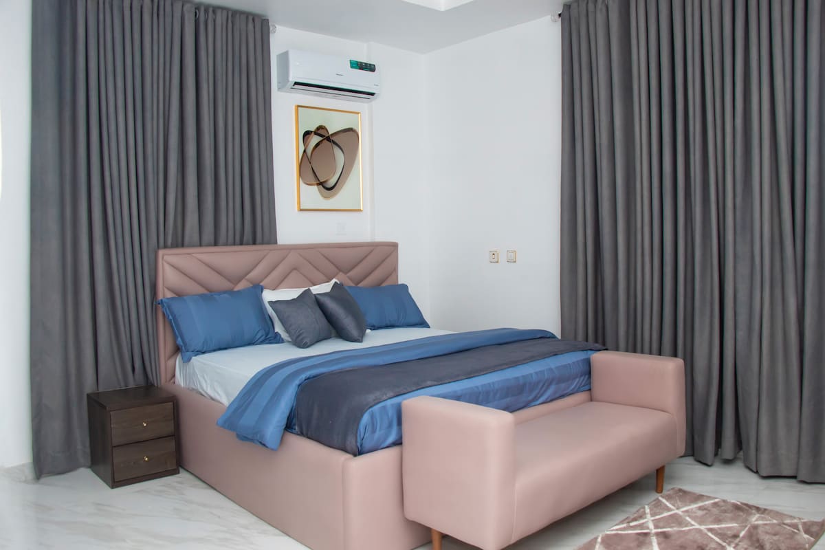 A sleeping area features a spacious bed dressed in soft blue and gray linens, complemented by decorative pillows. A pink settee is positioned at the foot of the bed. Floor-to-ceiling gray curtains frame a window, while a minimalist artwork is displayed above the bed.