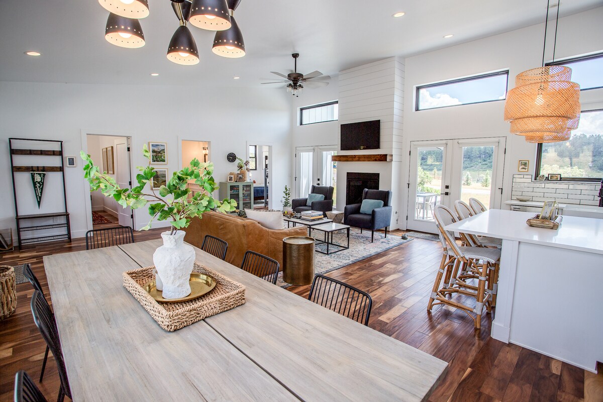 An open-concept living area features a large dining table with black chairs, a cozy living space with a brown sectional sofa, and a sleek kitchen island. Natural light fills the room through multiple windows, complementing the modern mountain decor and inviting ambiance.